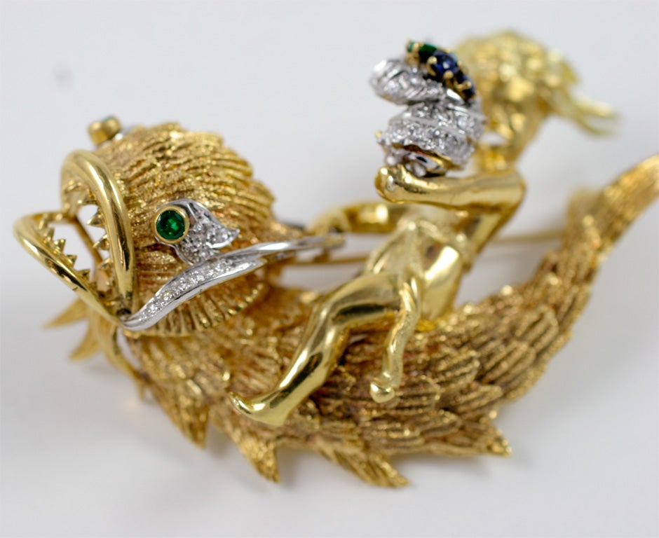 Highly detailed and ornate brooch of a “golden” boy carrying a torch, encrusted with pave diamonds, sapphires, and emeralds, holding on to an emerald-eyed dolphin with a diamond leash. What is more intriguing about this brooch is the symbolism