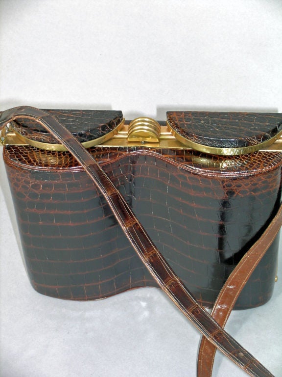 Dark brown alligator skin accented by a warm brass frame makes this shoulder bag shaped like a binocular case a true conversation piece.<br />
<br />
The bag has a double curve on the front section imitating binoculars.  This opens to reveal the