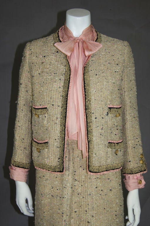Three piece beige iconic 1961 Chanel wool suit. Baby pink silk smocked blouse with cap sleeves and tie in front.  Skirt has front flap zip and is paneled all the way around. The jacket has brocade detail around edges and faux blouse cuffs with cuff