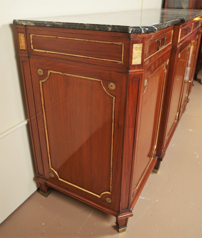 A grand mahogany parcel-gilt sideboard with a beveled dark gray and white vein marble, atop four narrow drawers over four matching cabinet doors, standing on squared bulbous feet and bronze casts. Stamped Jansen (underneath marble). Both front and