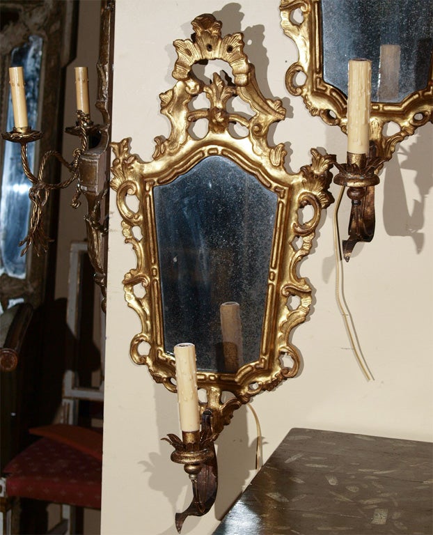 Pair of one arm Italian mirrored sconces in an elaborately carved and gilded wood frame. US wired