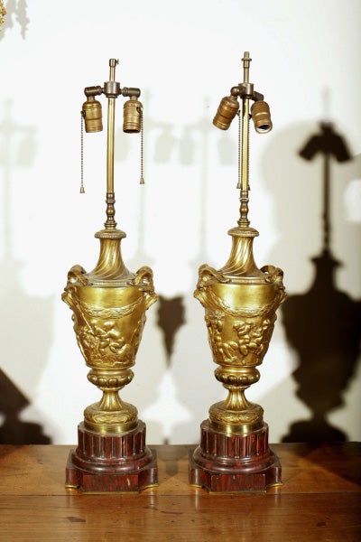 The  gilt bronze lamps on dark red marble fluted bases, each decorated with putti, swags and masks with ram type horns, each signed  CLODION (CLAUDE MICHEL, 1738-1814). in need of rewiring.