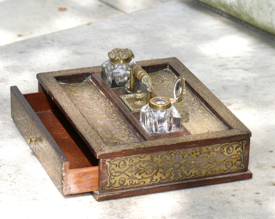 A brass inlaid English ink-well, supporting two chrystal ink bottles with decorated bronze lids. Having a drawer in front and a stamp apartment in the middle between the ink bottles.