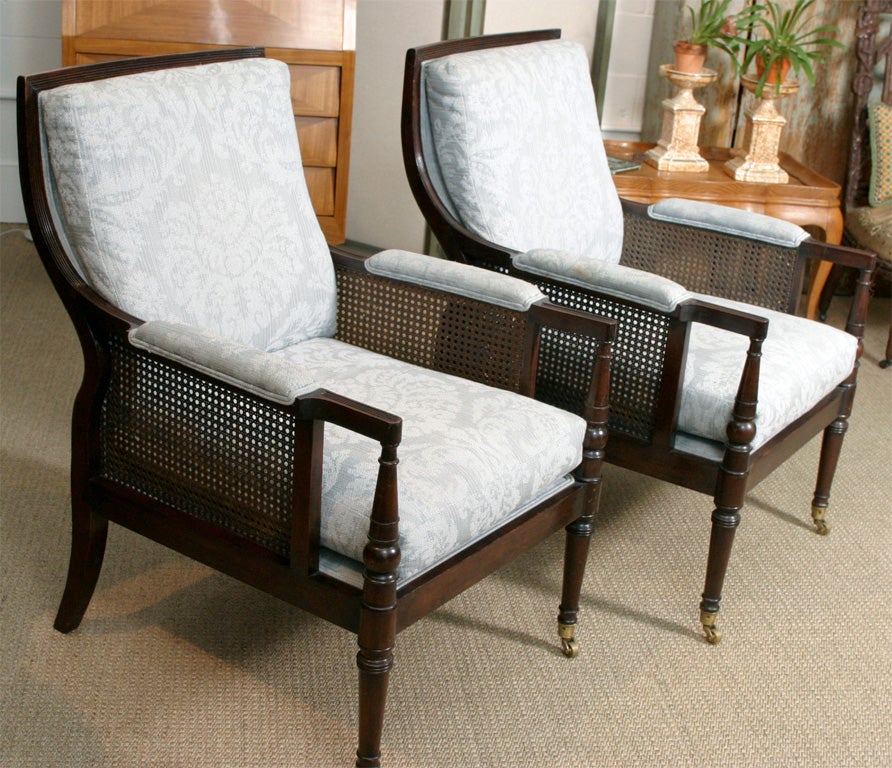 A very handsome pair of English mahogany upholstered chairs with caned sides and a contoured back. The front legs are capped in solid brass casters. These chairs are very comfortable and stylish.