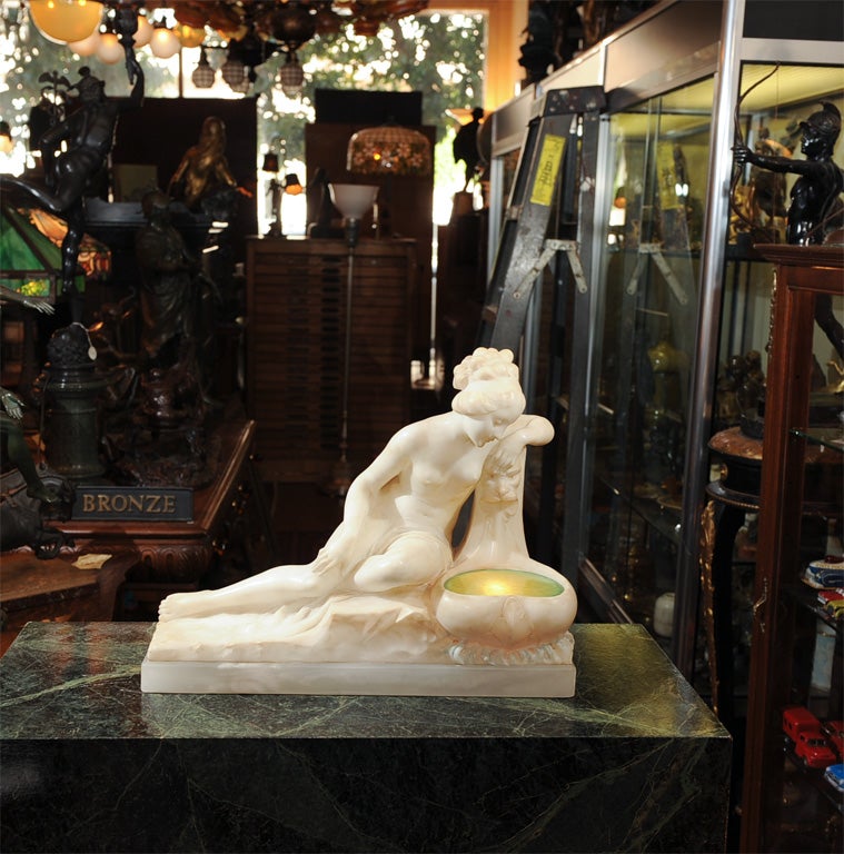 Alabaster figural lamp of a nude looking into a fountain.  This beautifully carved woman is resting on a naturalistic base and peering into a fountain that has a beautiful piece of green glass which lights up.  Her left arm rests on a carved