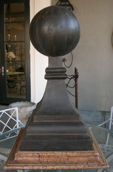 A boldly-scaled French zinc melon-form roof finial; capped by a large melon-form sphere above a splayed body over a stepped base; resting on a later faux marble painted wooden plinth