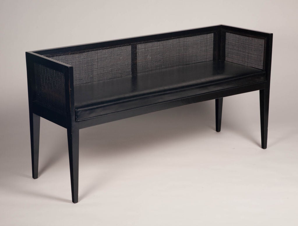 A long, slender bench with black leather upholstered seat, black painted caned back and sides, all resting on four square tapered legs, the whole finished in ebonized wood. Seat height is 19 1/2 inches. By Baker. American, circa 1960.