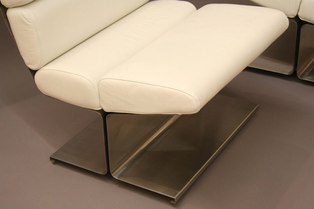 Pair of lounge chairs in brushed steel and leather, the steel frame in two parts forming the back and seat meeting to form a central support and spreading foot, the cushions in white leather