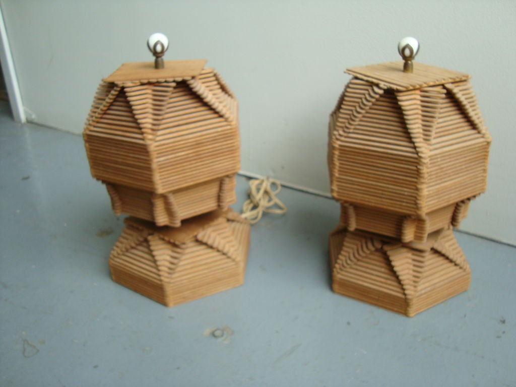 A hand crafted pair of popsicle stick lamp bases and shades constructed entirely of Popsicle sticks in a rotating pattern topped with ivory colored and brass finials.