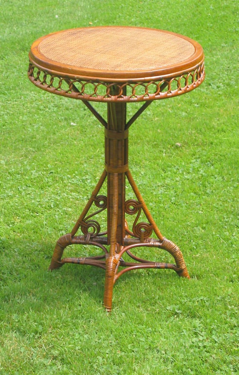 Early round wicker occasional table made of rattan, having top surface covered in simple pattern of pressed cane. Chain-looped apron and S-scrolls at base. Square pedestal post supported on four splayed legs joined by double stretchers.