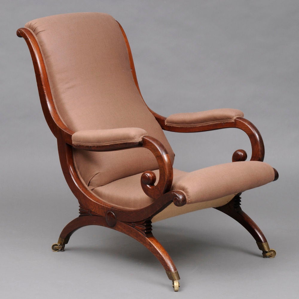 Regency mahogany X-frame, sleigh-shaped library armchair with arched leg and arm supports centered by roundels and carved stylized flower forms, scrolled and padded arms, raised on brass casters. Upholstered in taupe-colored fabric.