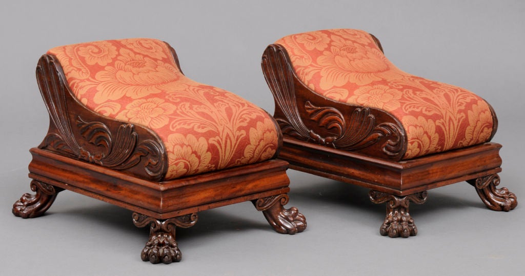 Pair of Regency mahogany footstools, the shaped padded top with carved sides of leafy scrolled pattern on molded frame, raised on carved paw feet. Upholstered in a damask fabric.