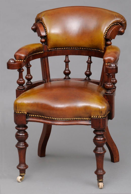 Victorian mahogany framed desk chair with scrolled back rest, turned arm and back supports, outward-scrolled hand rests, serpentine shaped seat, turned front legs on porcelain casters, outward splayed back legs. Upholstered in vellum-colored leather