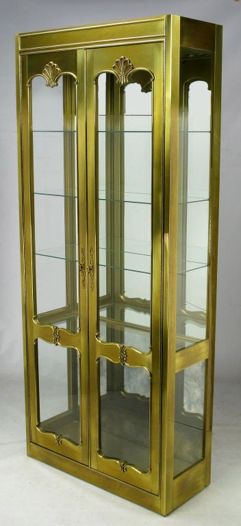 Elegant French Regency inspired brass display cabinet by Mastercraft. A pair of cased brass doors with shell and cartouche detailing, open to four glass shelves with room for another in the lower section. Venetian mirror back adds depth to the