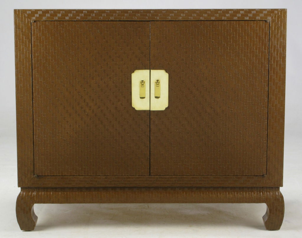 This chocolate brown lacquered grass cloth cabinet is from Baker's Far East Collection.  Features Asian style turned legs, and large brass escutcheons backing solid brass drop pulls. The lacquered grass cloth is in amazing like-new condition and is