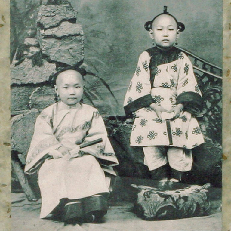 Vintage print of two young Chinese children in traditional dress, seated outside in a garden. Vintage Chinese postage stamp is affixed to upper right hand corner of image. Photo is backed with layers of textural rice papers. Wood frame has