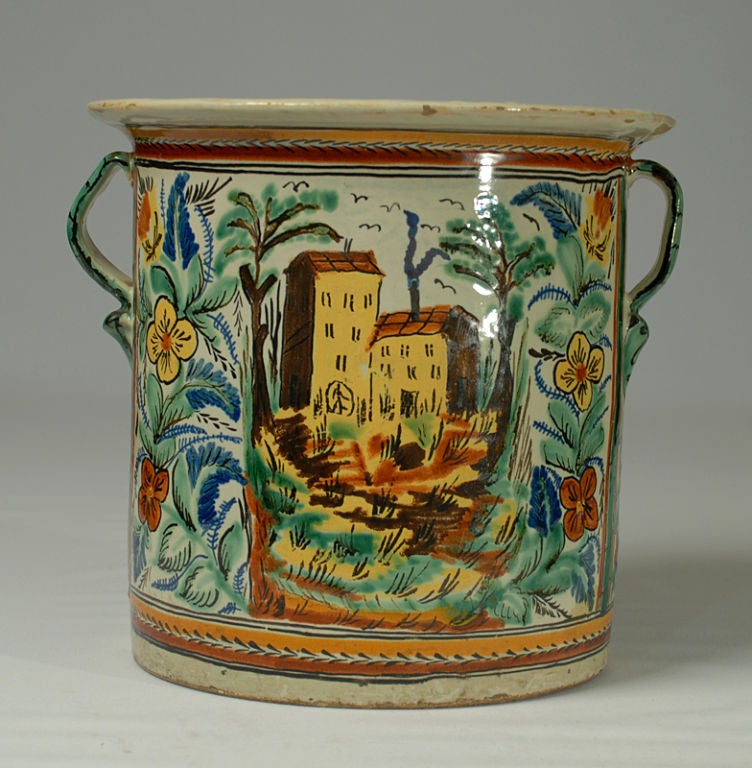 A good, large early 19th century Mexican talavera handled 'vasija' with a village scene surrounded by foliate patterns in green, yellow, dark brown, red and cobalt. The small pitcher is shown for scale only and is not part of this listing. <br