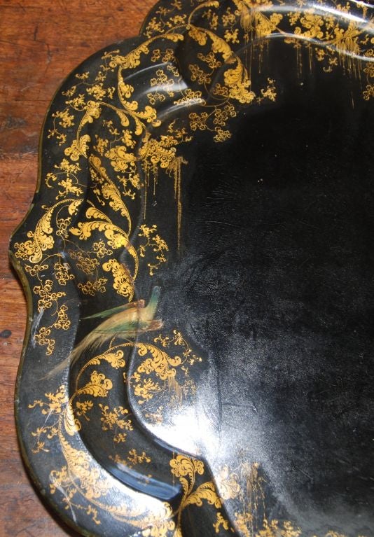 A large papier mache black lacquer tray with hand painted gold leaf floral decoration around the edge with birds. It is a delicately detailed English 19th century tray with a generous and deeply scalloped edging.