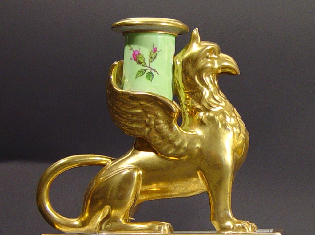 Each candlestick is modelled as a gilt griffin supporting a candleholder between its wings.  Each griffin is seated on a high rectangular pale green ground painted plinth reserved on the front and back with finely painted bouquets of flowers with
