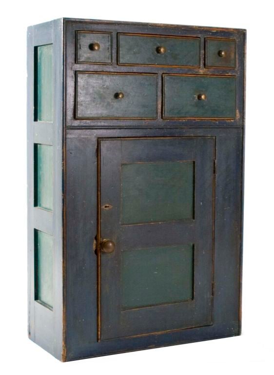 This hanging cupboard has a three-over two-drawer arrangement over a paneled door with original escutcheon and pulls. The cupboard is painted an old dark-blue paint with green painted drawers. It has three sunken panels on each of the sides.