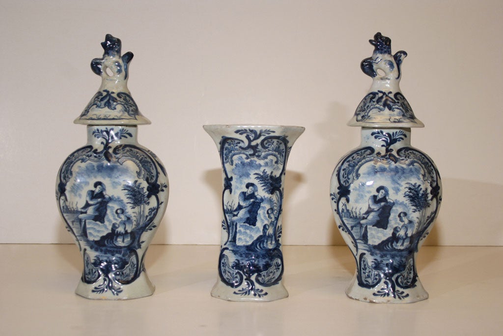 COMPRISED OF TWO OCTOGONAL COVERED BALUSTER VASES AND ONE OCTOGONAL BEAKER, EACH PAINTED WITH A SCENE DEPICTING A MAN AND A WOMAN HOLDING A BABY AND A WINDMILL IN THE DISTANCE RESERVED WITHIN SCROLLED FOLIATE BORDERS, WITH AXE FACTORY MARK.
