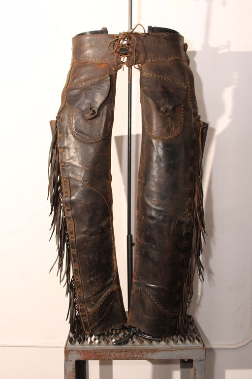 Fantastic pair of Vintage Rifle-Leg Western Chaps with intricate leather tooling and stitching throughout; hand-cut fringe at each leg and a scalloped lace-up belt.  The original custom telescoping  stand is decorated with a horseshoe base.