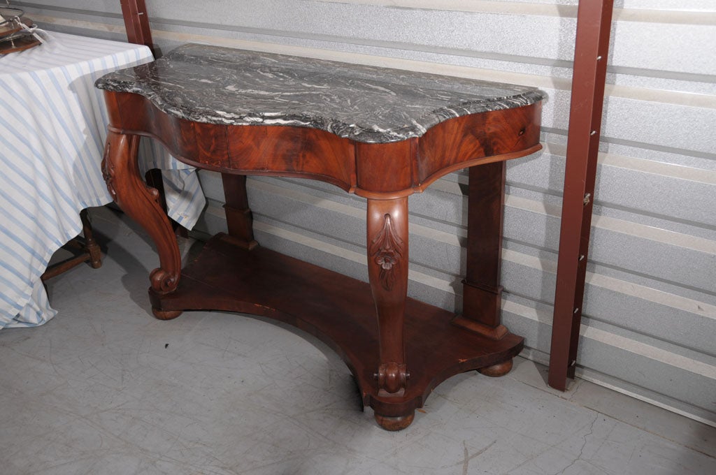 A fine Italian serpentine console table featuring a moulded-edge top of gray figured marble set upon a frame of mahogany. 
The frame - a frieze of flame-cut mahogany above two scroll-carved front supports adjoined to a shaped base and resting on bun