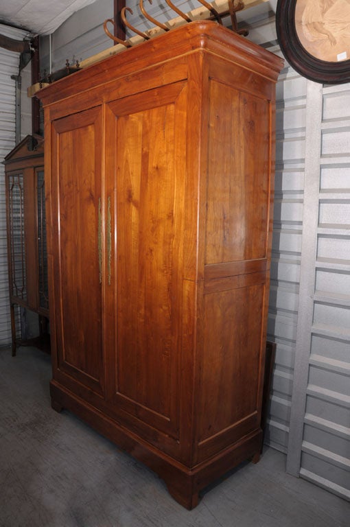 A French armoire (or bookcase display cabinet) of cherry wood in the Louis Philippe style featuring a moulded crown above two framed doors (each with decorative brass escutcheons), enclosing a cupboard with three shelves, and resting on formed