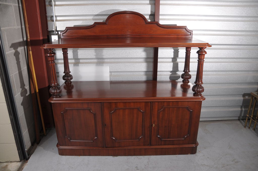 A fine English serving console or sideboard of mahogany, featuring a moulded-edge top tier with a shaped back, set upon four ring-turned column supports resting on a bottom tier cabinet on plinth.

The cabinet featuring a moulded-edge top above