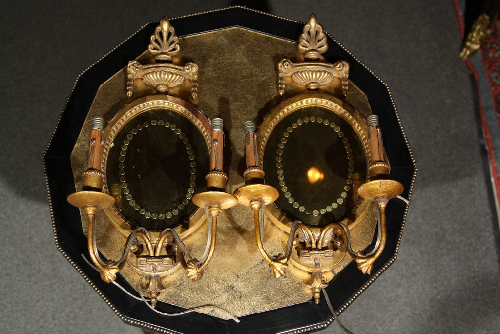 Pair of French gilded two-arm mirrored-back wall sconces, the beaded oval frame decorated with urn crest.
