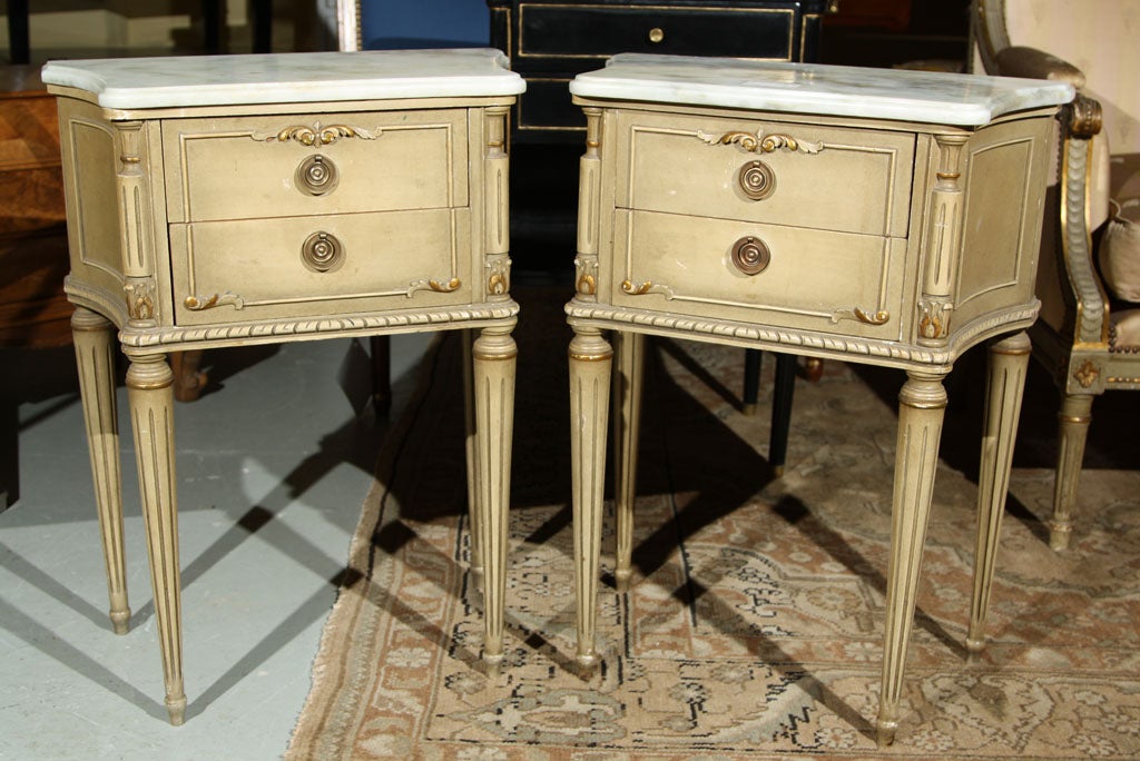 Pair of nightstands, in the style of Louis XVI, ivory painted throughout with parcel-gilt highlights, the beveled white marble top over a small case of two drawers, standing on tapering circular legs. Stamped Jansen.
