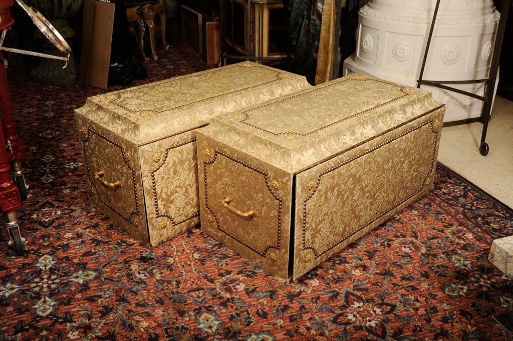 This pair of vintage cartonnnier storage chests feature luxurious golden colored upholstery decorated with nail heads and rosettes, and metal side handles. Each has a front panel which swing upward to reveal nine vertical storage compartments, ideal