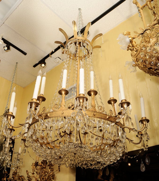 This antique neoclassical bronze chandelier features a circular frame with lower canopy of faceted crystal chains and eleven candles, and outer ring of twelve candle arms. A central stem connects the lower frame with an upper ring decorated with