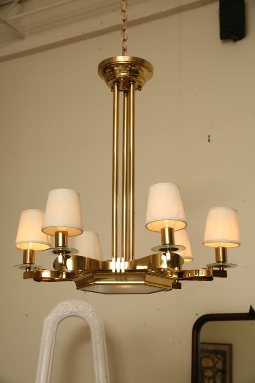 This elegant and classic French vintage 1940s six light polished brass chandelier is greatly inspired and in the style of Gilbert Poillerat. It has glass discs and linen shades, suspended from four columns and having a cut glass centre plate
with a