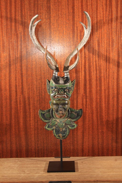 Old wood carving of a Burmese Nat (celestial being or spirit figure), depicting a human figure with head thrust forward and wearing a demon mask head dress mounted with antlers. The over all figure painted in green pigments with silver highlights