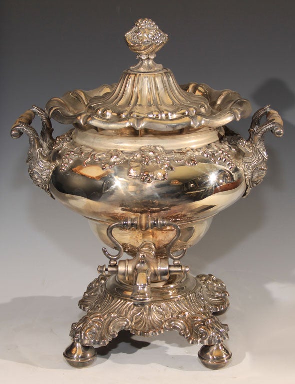 An English Sheffield plate hot water or tea urn, having vine leaf and grape motifs. The inverted pear-shape urn has two wooden handles on each side flanked by leaf and scrolled cartouches and a spout in the front. The urn sits on a shell, acanthus,