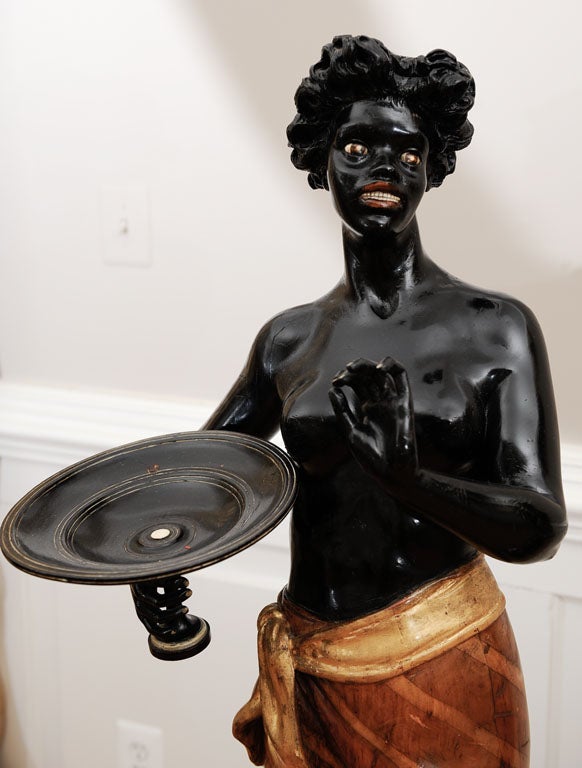 Pair of Blackamoors with Mother of Pearl eyes, teeth, nails, gold leaf draped sheafs, both holding tazzas. Female: wearing sarong skirt. Natural wood with gilt sash, holding tazza in right hand. 61