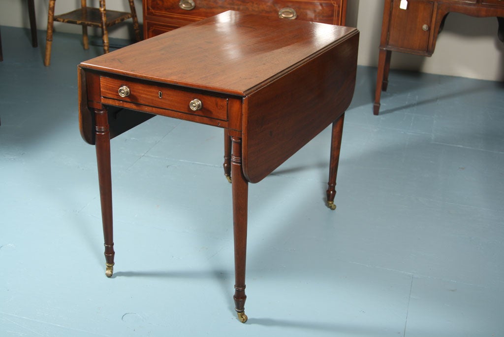English mahogany Pembroke table on turned legs ending in brass casters. Rectangular top with rounded corners; single pine-lined drawer with brass knobs.