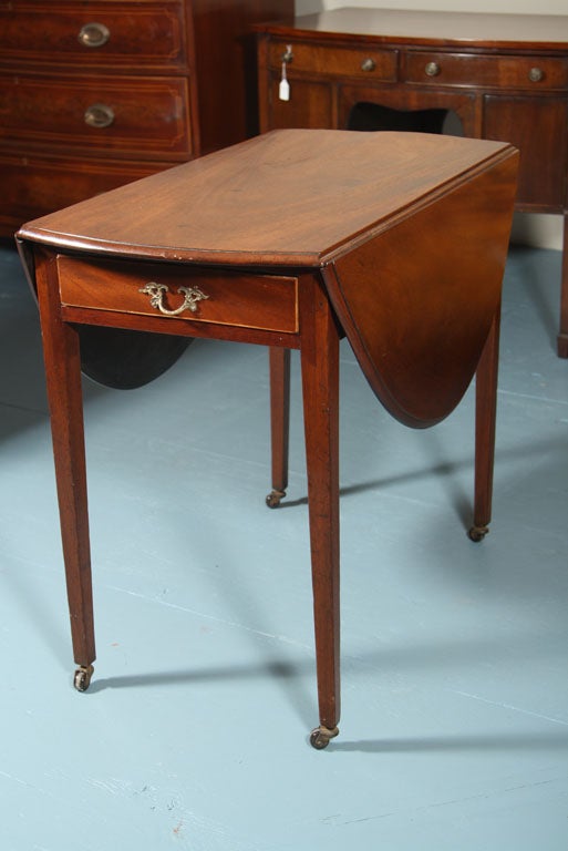 English, solid mahogany oval Pembroke table on tapered legs ending ceramic caster with brass fittings. Features include a beautifully figured mahogany top and satinwood stringing on the drawer front, which has a replaced brass pull. In a testament