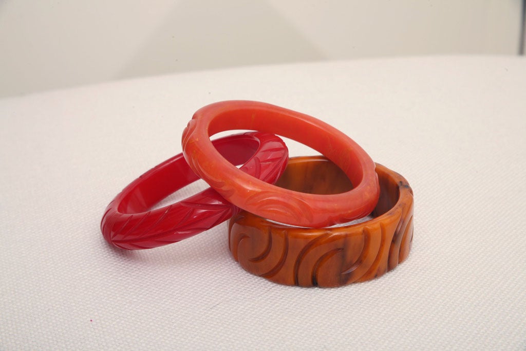 Il set di 3 braccialetti in bachelite art deco autentico vintage intagliato è nelle tonalità del rosso, dell'arancio ambrato e del caramello. Sono tutti intagliati a mano e quello al caramello arancione è un bracciale più spesso con piccole volute