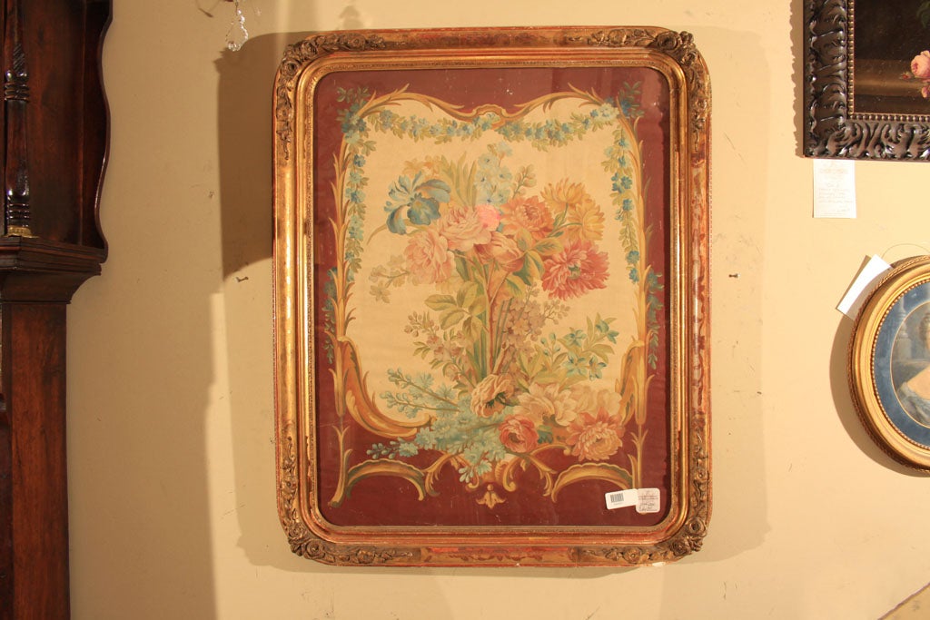 Aubusson style painted flowers and swags in shades of pastel blue, red, yellow and green on cream colored ground framed by a cartouche on dark red ground and border. Rectangular frame with round corners carved with flowerheads and foliage, gilded