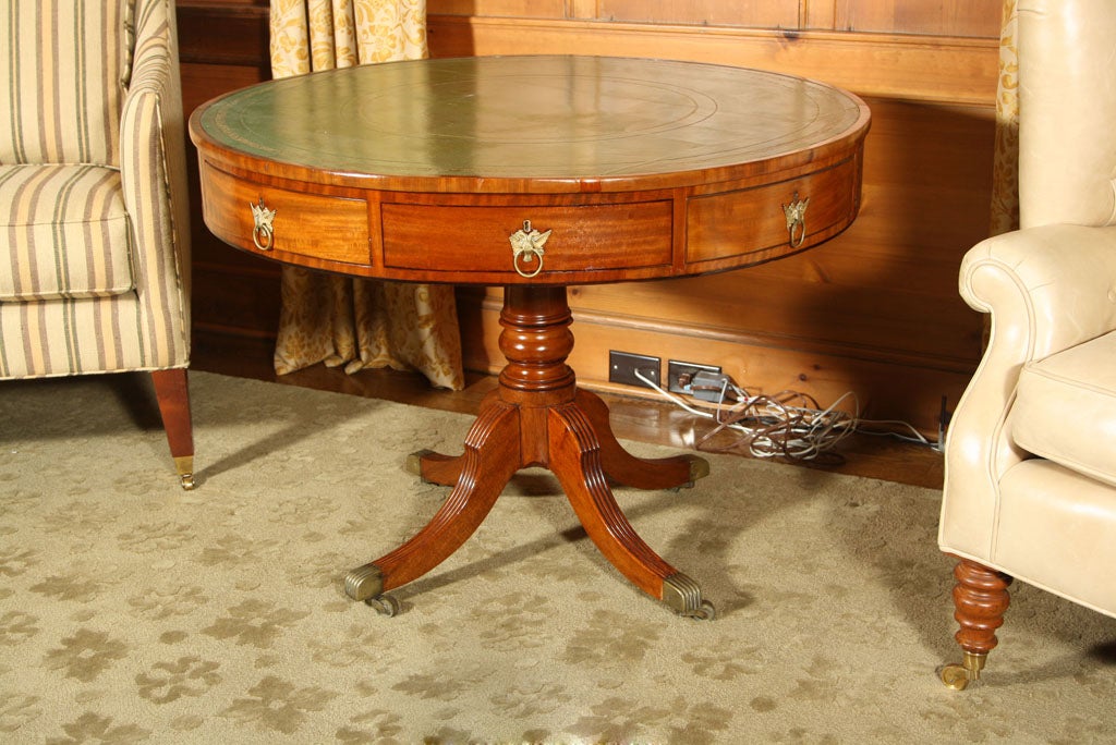 DRUM TABLE NEATLY EXECUTED IN RICHLY GRAINED MAHOGANY. THE TOOLED GREEN LEATHER TOP SURROUNDED BY A CROSSBANDED BORDER AND EDGE. THE FRIEZE WITH DRAWERS ALL AROUND. THE PEDIATAL SUPPORT WITH FOUR SPLAYED AND REEDED LEGS ENDING IN BRASS CASTERS WHICH