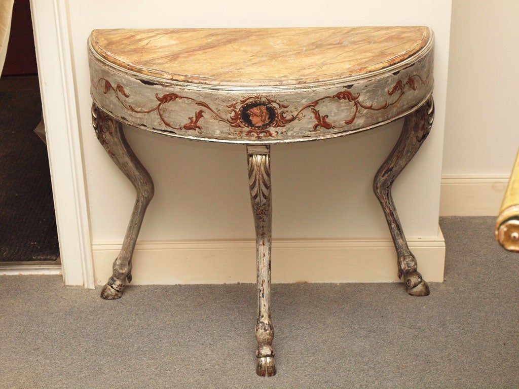 Delicate silver gilt wood and painted demilune with zoomorphic legs terminating in pied de biche. The front of the demilune is adorned with a neoclassical painted head and flanking  scroll ornamentation.  The top is faux marbre and is removable. 