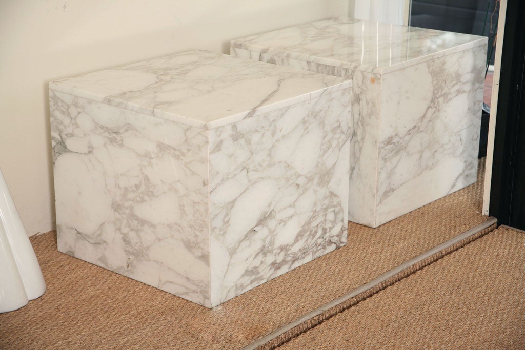 Two tables fashioned of solid carrara marble slabs. Four sides of equal proportion at 20  inches high with the tops resting on those sides at 24 inches square. Can be used in a myriad of ways; side tables next to sofa or across from one another next