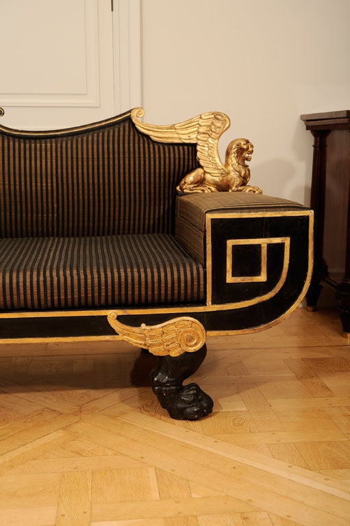 The upholstered back with scrolling toprail terminating in uprights in the form of seated winged lions, above upholstered armrests opening to a plain apartment, the upholsted seat above a shaped border terminating in a Greek key scroll, the whole