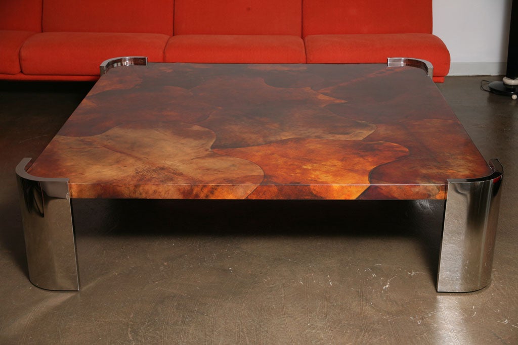 Six foot square coffee table attributed to Karl Springer covered in hues of orange and brown goat skin. Four curved columnar steel legs in mirrored stainless steel.