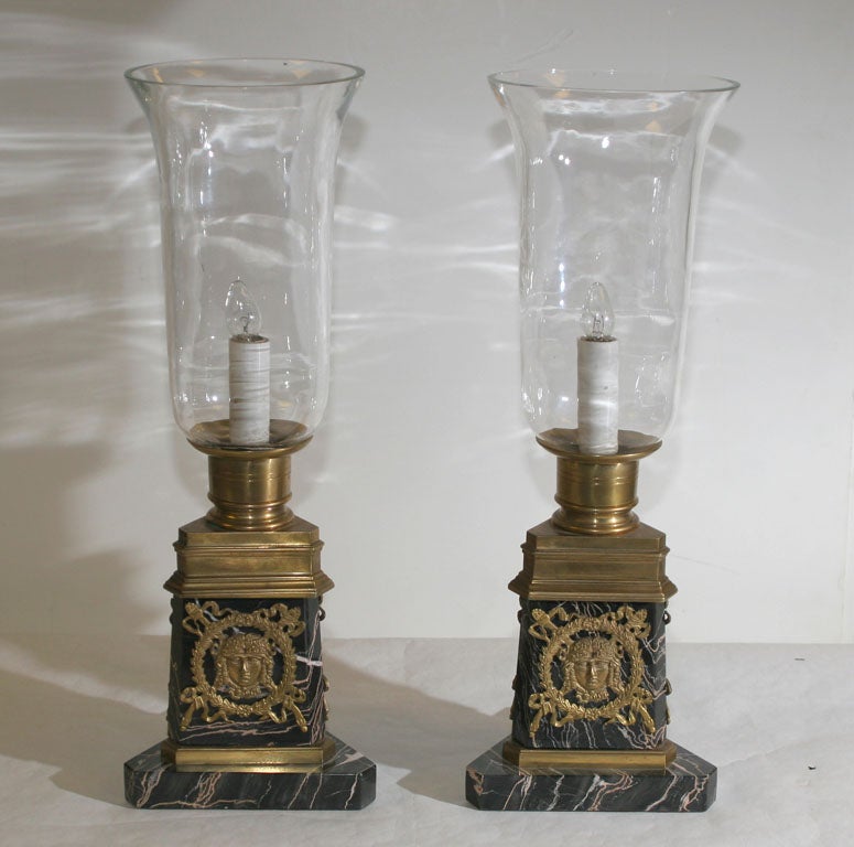 EACH WITH A GLASS SHADE RESTING ON A CIRCULAR NOZZLE ABOVE A CANTED TRIANGULAR PLINTH ENDING ON A CONFORMING BASE, THE PLINTH APPLIED WITH GILT-BRONZE MASKS. ELECTRIFIED.