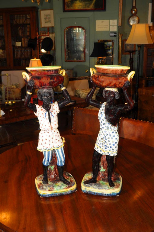 A pair of Majolica black-a-moors featuring a man and woman in exotic, patterned dress holding pots above their heads, standing on bases in front of spill vases moulded as tree trunks.<br />
<br />
They are attributed to Thomas Sergent, a