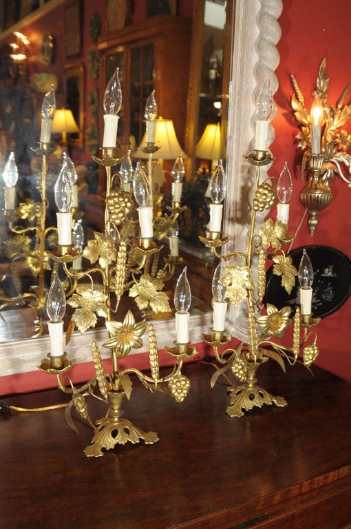 A lovely pair of French girandoles (five-light candelabras) of gilt metal embellished with a design of flowers, grape leaves, grape clusters, and wheat stalks. 
Each girandole featuring five candle lights (with leaf-design bobeches) on stem-like