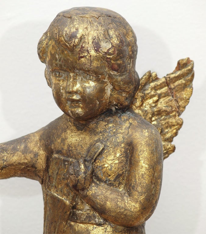Carved giltwood sculpture of a cherub standing on a rock.  It is draped in a cloth with a quiver of arrows slung across its' shoulder and is holding a metal flower.  It is mounted on a round stand.